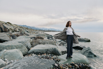 Young woman stands on rocky beach wearing coat and white shirt. Calm sea and cloudy sky in background create peaceful atmosphere. Concept for freedom, nature, and contemplation. © SHOTPRIME STUDIO