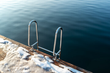 A stainless steel ladder descends from a snow-covered wooden dock into calm dark water. Winter swimming concept, minimal waterside detail, and cold season outdoor background. © Volha