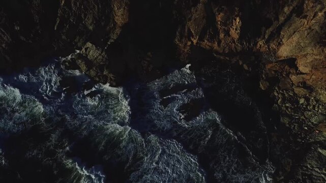 Dramatic aerial view of ocean waves crashing against rugged, dark cliffs at sunset