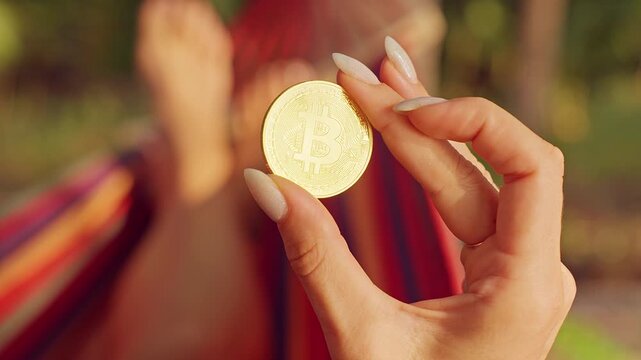 Close Up First Person View of Golden Bitcoin Coin Held Between Fingers With Hammock and Tropical Background Showing Financial Freedom and Digital Asset Value