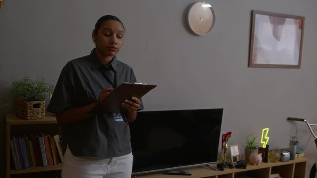 Handheld shot of young Black female social worker taking notes while assessing living conditions during home visit to teenage boy, lying on floor in messy bedroom in relaxed pose