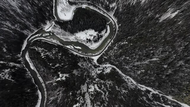 FPV drone top-down flight over Cheile Zugrenilor island, Romania, showing winding mountain road and river curves through forested valley with patches of snow in winter.