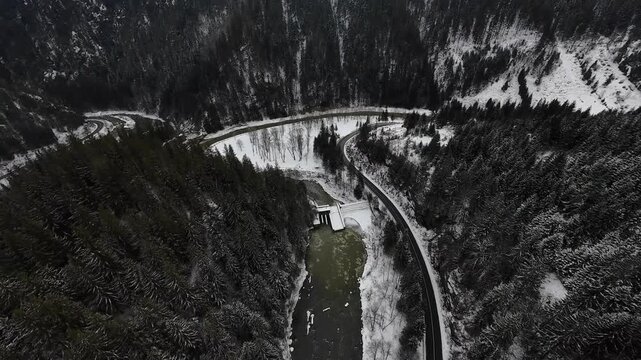 FPV drone top-down flight over Cheile Zugrenilor island, Romania, showing winding mountain road and river curves through forested valley with patches of snow in winter.