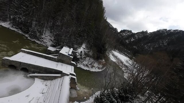 FPV drone top-down flight over Cheile Zugrenilor island, Romania, showing winding mountain road and river curves through forested valley with patches of snow in winter.