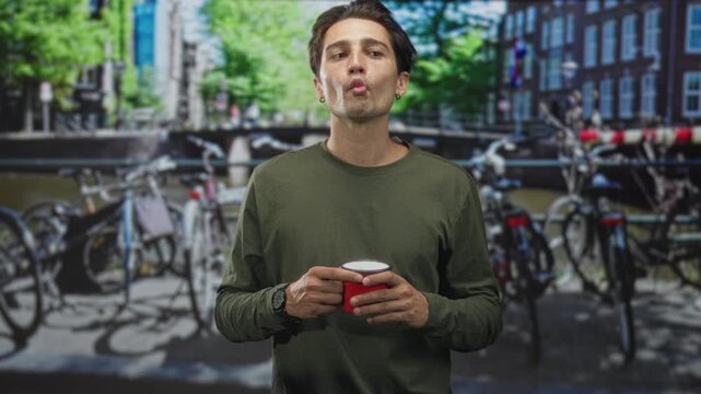 Man holding red cup purses lips on street by canal with bicycles and bridge, wearing green shirt, earring and watch for a casual moment; playful pause.