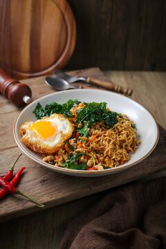 Spicy Thai Stir-fried Instant Noodles (Mama Pad Kra Pao) with Minced Pork, Holy Basil, and a Crispy Fried Egg on a Rustic Wooden Table.