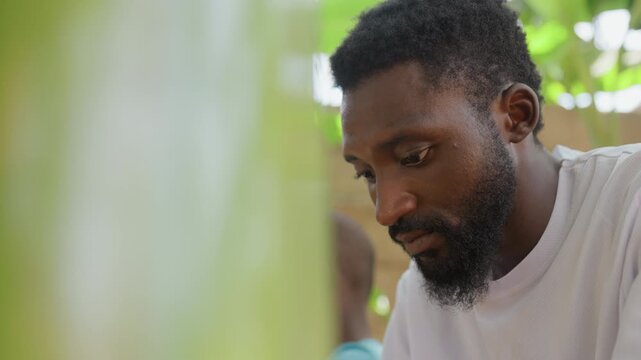 Closeup man portrait in garden quiet outdoor scene showing contemplative adult with beard and downward gaze, blurred leaf foreground, soft sunlight, candid moment suggesting pause and reflection,