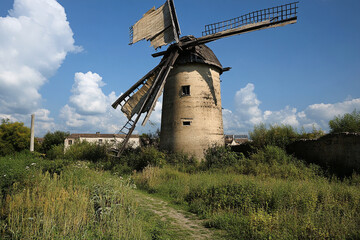 Fototapeta premium Abandoned windmill with broken sails surrounded by tall grass and wild vegetation on a sunny day in a rural landscape