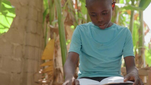 Youth engaged in learning on shaded porch. Young boy attentively examining pages beneath natural sunlight. Boy deeply focused on his book while seated in peaceful garden environment