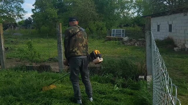 Man using a string trimmer to cut grass in a backyard on a sunny day in the countryside