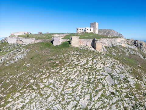 Aerial view of the Castillo de la Estrella perched atop a rugged hill, its ancient stone walls glowing warmly under the clear blue sky, Teba, Andalusia, Spain.