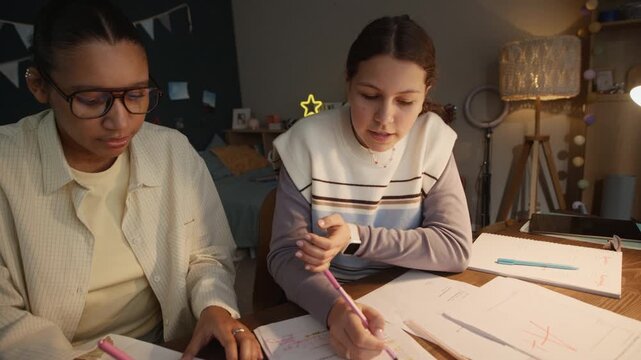 Tutor and student sitting at desk, teen girl doing math tasks and communicating with young woman