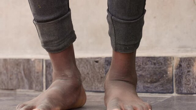 Barefoot black child feet on tiles, sportready stance with dusty ankles and sandy soles, rolled dark leggings, closeup texture of skin and detail of toes, urban threshold and muted wall, quiet playful