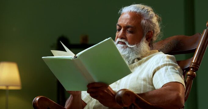 Indian mid age bearded man falls asleep while reading book novel on relaxing chair at home, stylish mature male with closed eyes holding printed copy in modern interior leisure lifestyle setting