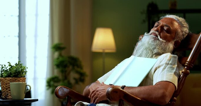 Indian mid age bearded man falls asleep while reading book novel on relaxing chair at home, stylish mature male with closed eyes holding printed copy in modern interior leisure lifestyle setting