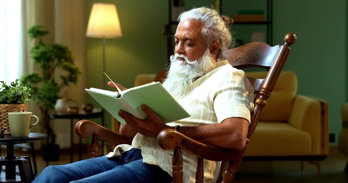 Indian mid age bearded man reading book novel on relaxing chair at home, stylish mature male enjoying peaceful leisure time with printed copy in modern interior lifestyle setting