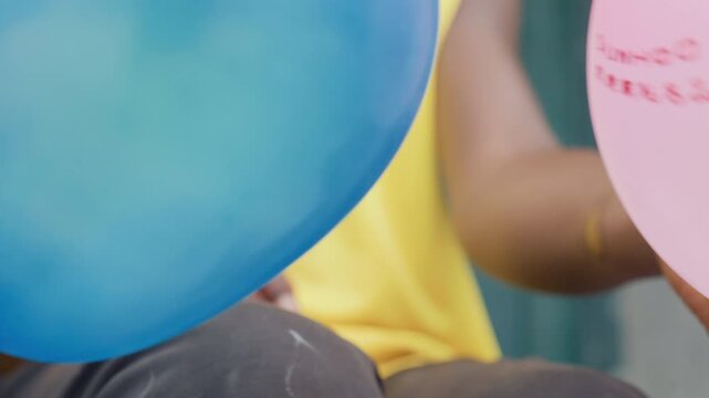 Blue balloon in hand closeup, bright color against yellow shirt, steady fingers tying knot, glossy latex sheen, playful movement, focused concentration, lap and casual outdoor scene