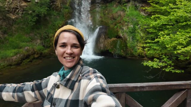 Female travel blogger filming selfie video near Veliki Buk waterfall in eastern Serbia. Smiling woman presenting scenic cascade and emerald pool in lush natural environment
