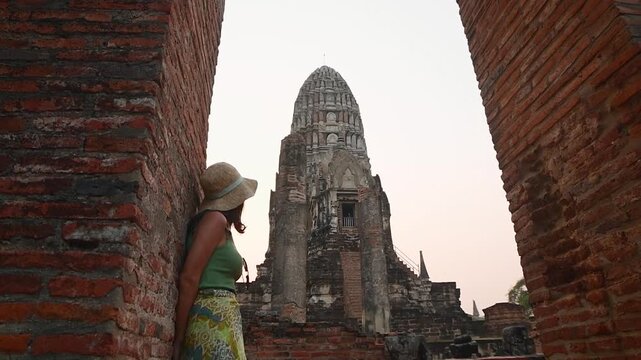 Ayutthaya, Bangkok -15th march, 2024: Female traveler in hat stands between brick walls at Wat Ratchaburana, Ayutthaya, watching ancient prang tower at sunset, Thailand heritage tourism concept