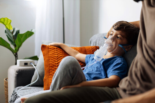 Young child receiving nebulizer treatment at home while sitting on sofa, representing pediatric respiratory care, asthma management and family health routine.
