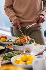 African American male carving roasted chicken with knife and fork at dining table in tan sweater