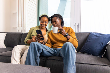 African American couple sitting on dark gray sofa in living room, sharing smartphone and mug