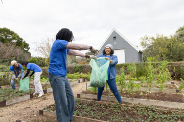 Diverse volunteers exchanging green compost bag in community garden by wooden beds wearing gloves
