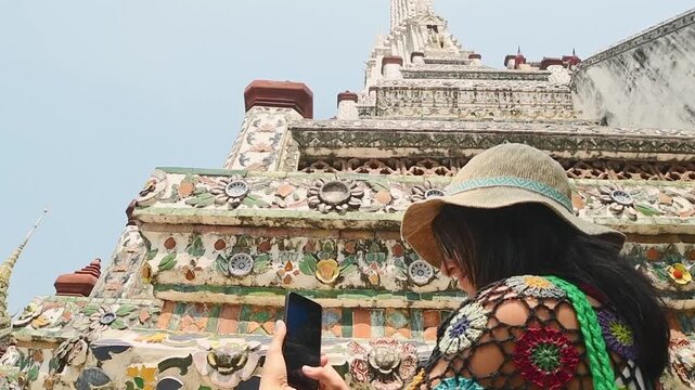 Bangkok, Thailand - 15th february, 2024: Bangkok Wat Arun Temple of Dawn, tourist woman in straw hat use smart phone to photograph colorful ceramic mosaic details, Thai Buddhist landmark