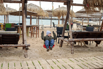 Happy little boy in winter clothes swinging on playground swing at sandy beach cafe area near the sea, enjoying outdoor childhood moment on cool cloudy day by the coast