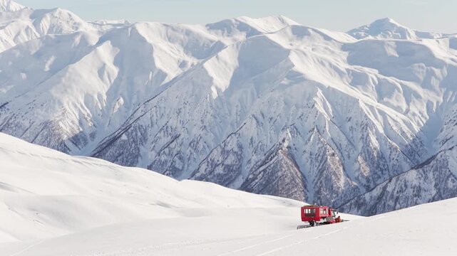 Aerial view Red snowcat drops riders on high alpine ridge, snowboarders prepare for freeride descent, vast Caucasus mountain range background, winter adventure tourism concept