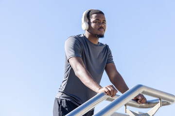 Fototapeta premium African American man leaning on metal stair rail in workout clothes, listening with white headset