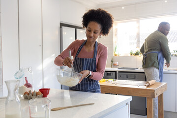 African American couple in striped aprons preparing batter in kitchen, woman whisking in glass bowl