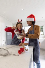 Fotobehang Koken Diverse couple wearing aprons and holiday hats pouring glass measuring cup into bowl at kitchen  © wavebreak3