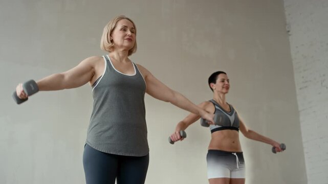 Low angle shot of two middle-aged women in sportswear doing lateral raises with light weight dumbbells during group fitness training