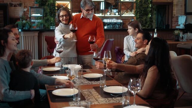 Happy senior couple giving a speech and toast during anniversary celebration with multi-generational family in modern restaurant. Emotional festive family moment.