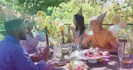 Clinking glasses, four adults wearing cone hats chatting on wooden deck with gold 30 balloons, cake