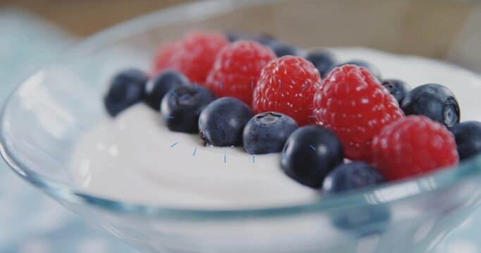 Showing glass bowl with white yogurt, blueberries, raspberries on pale cloth, rim reflecting light