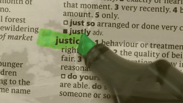 Gloved hand entering right touching page, green marker laying strip highlighting justice dictionary