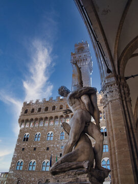 Low-angle view of the dramatic marble sculpture 'Hercules and Cacus' in Piazza della Signoria, framed by a loggia arch and the towering Palazzo Vecchio.