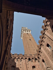 Obraz premium Dramatic low-angle view of the towering red-brick Torre del Mangia framed by the medieval architecture of the Palazzo Pubblico inner courtyard in Piazza del Campo, Siena.