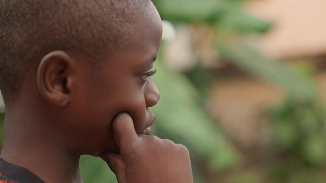 Bright young boy looks with gentle smile. Youthful boy with bashful expression among leafy trees in warm hues. Inquisitive young child gazes softly with shy smile amid lush greenery in warm colors
