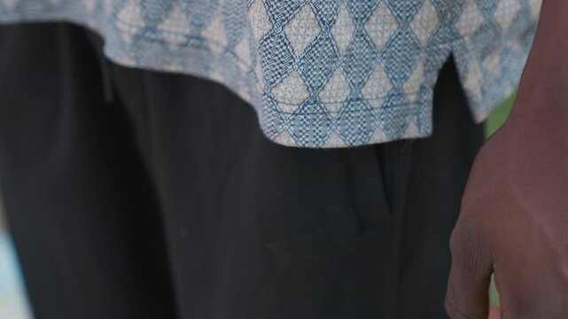Black man resting hand in pocket, patterned shirt and black sport shorts. Closeup outdoor portrait shows textured fabric, dark skin arm, relaxed stance during training break, soft sunlight and urban