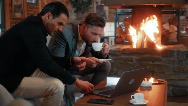 Two successful businessmen collaborating and negotiating while looking at laptop, sitting by fireplace in luxury hotel. Professional corporate meeting in elegant interior.