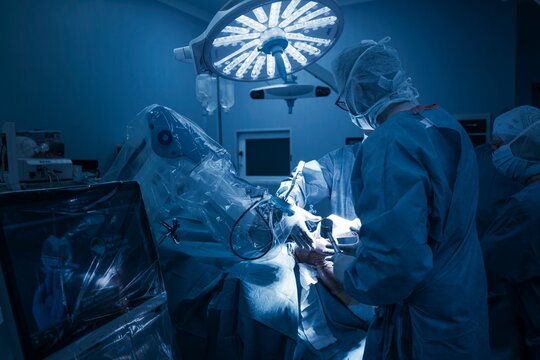 In a modern operating room, an orthopedic surgeon guides a robotic arm for knee replacement surgery. A touchscreen console shows 3D models and data for precise alignment