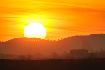 Fototapeta premium As the sun descends below the horizon, it casts a warm golden glow over rolling hills, painting the sky with shades of orange and yellow. A peaceful home rests quietly in the foreground
