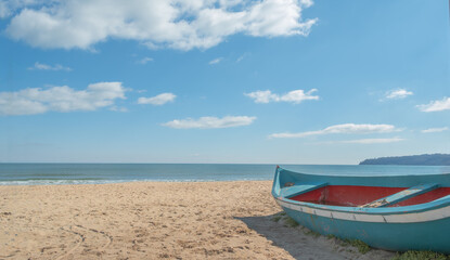 Obraz premium Rustic blue and red wooden fishing boat resting on a golden sandy beach. Scenic coastal view with calm sea, clear blue sky, and horizon in the background