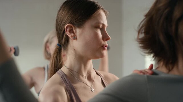 Close-up shot of young female instructor teaching middle-aged woman to do dumbbell wide arnold press during group fitness training