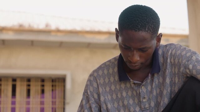 Young black man sitting on porch, looking down in contemplative mood closeup sequence shows patterned shirt, barred window with purple curtain, daylight exterior of modest home, pensive expression