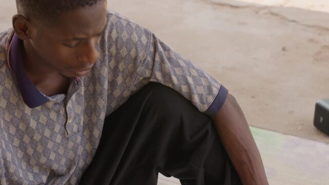 Young black man sitting on concrete, wearing patterned polo and black pants, head bowed in quiet contemplation, hands near knees, soft natural light, intimate documentary mood, cinematic close