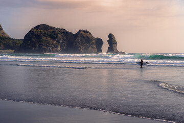 A surfer with a board walks into the ocean at Piha Beach, New Zealand, Auckland, with Lion Rock in...